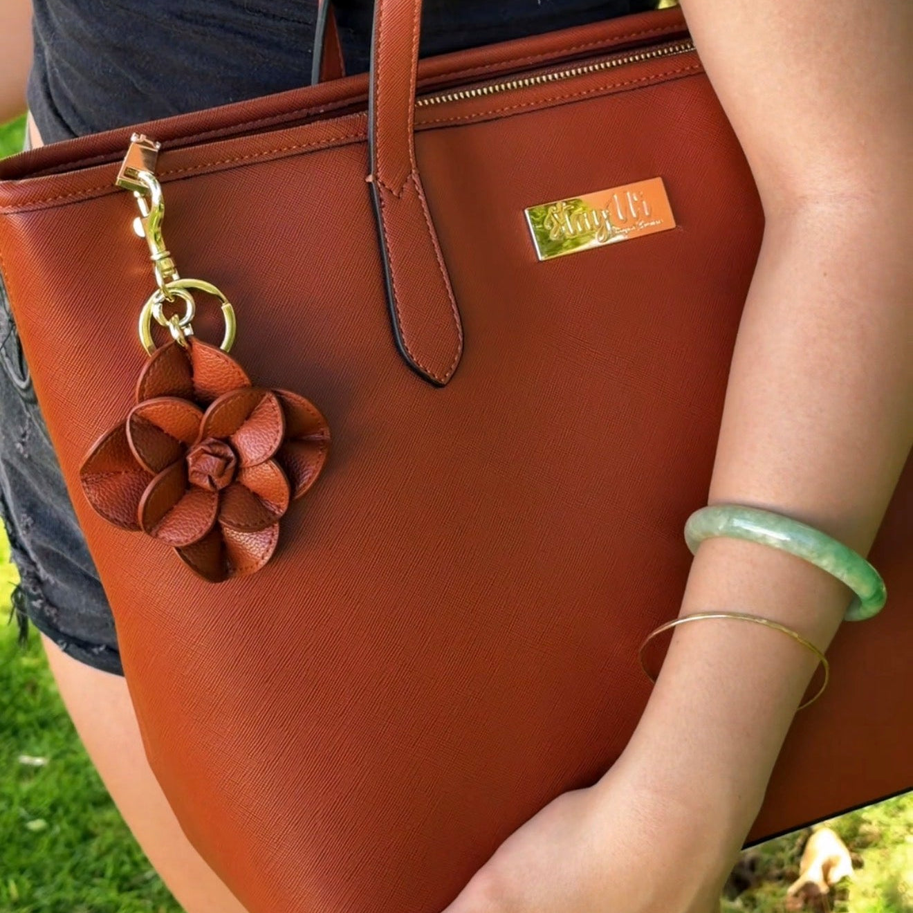 Brown leather tote bag held by a person with a decorative flower charm, outdoors.